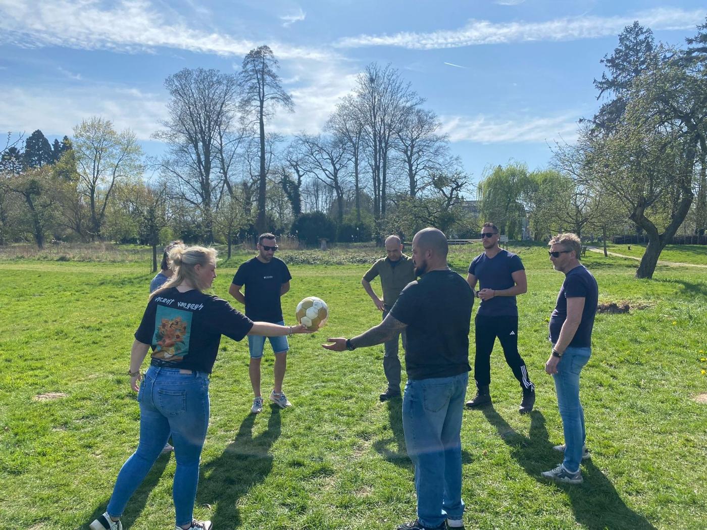 Een team staat in een kring op het gras en geeft een bal door tijdens een opdracht van Kennisgevers