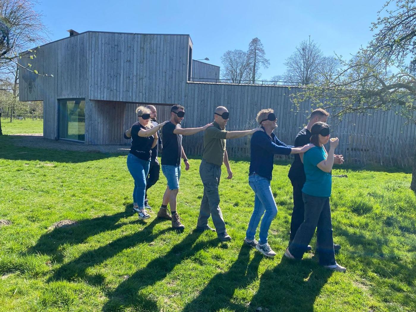 Teamleden lopen geblinddoekt achter elkaar, met een hand op de schouder van de voorganger, tijdens een teamdag van Kennisgevers